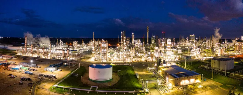 Refinería iluminada en hora azul con tanque de almacenamiento y antorchas